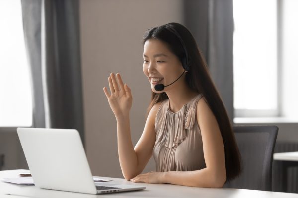 Smiling asian businesswoman wearing wireless headset waving hand doing video chat looking at laptop consulting customer sitting at workplace, female call center service support agent talking ; Shutterstock ID 1408485929; purchase_order: -; po: -; client: -; other: -